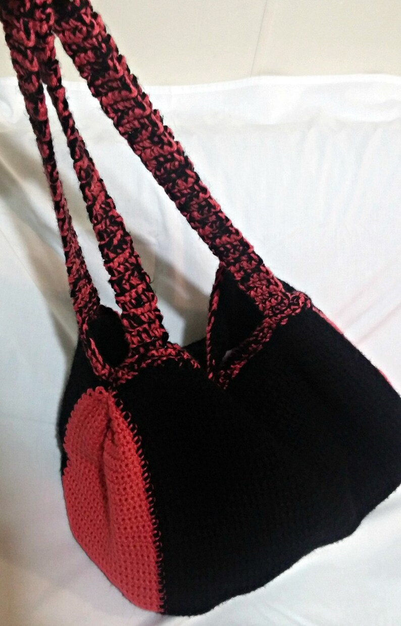 May include: A black and red crocheted tote bag with two straps. The bag is made with a textured stitch and has a heart-shaped design on the bottom.