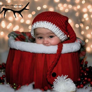 Puede incluir: Un bebé con un gorro de Papá Noel de crochet rojo y blanco con un largo pompón blanco, sentado en un cubo de madera rojo con ribete de piel blanca. El bebé mira a la cámara con una expresión seria. El fondo es una imagen borrosa de luces navideñas blancas.