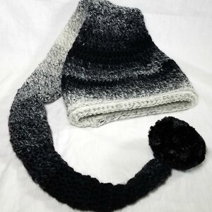 May include: A black and white ombre crocheted hat with a long tail and a black fur pom pom.