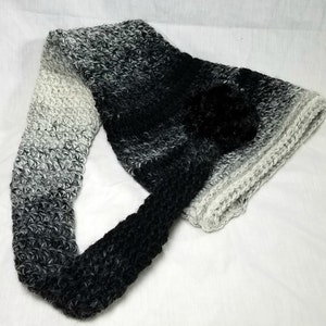 May include: A black and white ombre crocheted scarf with a black pom-pom.