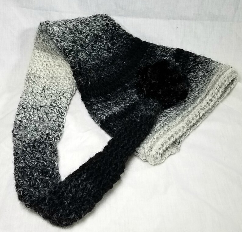May include: A black and white ombre crocheted scarf with a black pom-pom.
