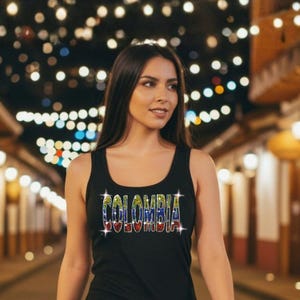 May include: A black tank top featuring the word COLOMBIA printed across the chest in a multi-colored, sparkling font that mimics the colors of the Colombian flag. The tank top has a scoop neckline and a sleeveless design, paired with blue jeans.