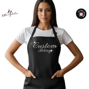 May include: A black apron with two front pockets and the text "Custom Bling" in silver script. The apron is worn over a white t-shirt. The apron has a halter neck and a simple design.