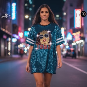 May include: A teal sequined t-shirt dress featuring a graphic of a face wearing sunglasses and the red text 1982. The sleeves have two white horizontal stripes. The garment is covered in shimmering sequins and has a round neckline.