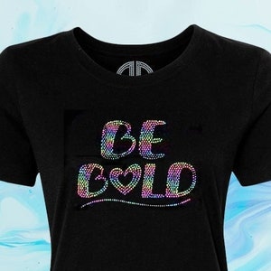 May include: Black t-shirt featuring a rainbow rhinestone design that reads "BE BOLD". The design includes a small heart in the word "BOLD". The text "Glossy Neon" is written in white on a black rectangle with the text "SAMPLE" in rainbow rhinestones below it. The text "By @Blingdots" is written in white at the bottom of the shirt.