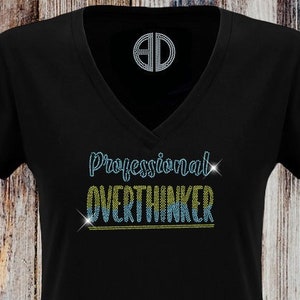 Könnte beinhalten: Schwarzes T-Shirt mit V-Ausschnitt und dem Text "Professional Overthinker" in blauen und gelben Strasssteinen. Der Text ist mit silbernen Strasssteinen umrandet. Das Shirt hat ein kleines Logo am Halsausschnitt, das "BD" sagt. Der untere Teil des Shirts hat den Text "By @Blingdots" in weiß.