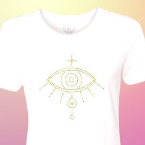 Könnte beinhalten: Weißes T-Shirt mit einem goldenen Augen-Design aus Strasssteinen. Das Auge hat einen kleinen Kreis in der Mitte und ist von Lichtstrahlen umgeben.