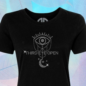 以下が含まれることがあります： 黒いTシャツに、ラインストーンで装飾された目のデザインと「THIRD EYE OPEN」の文字、その下に三日月が描かれています。「By @Blingdots」の文字がTシャツの下部に書かれています。