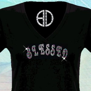 May include: Black v-neck t-shirt with the word "Blessed" spelled out in rhinestones. The rhinestones are multi-colored and sparkle. The shirt has a small logo on the neckline that says "BD". The text "By @Blingdots" is printed on the bottom of the shirt.