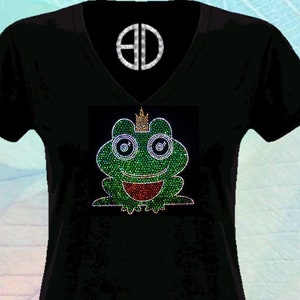 Może przedstawiać: Czarny T-shirt z zieloną żabą w złotej koronie, wykonaną z kryształków. Żaba ma duże, niebieskie oczy i czerwony pysk. Na dole koszulki znajduje się napis "By @Blingdots".