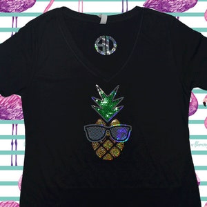 May include: Black t-shirt with a sparkly pineapple design wearing sunglasses. The pineapple is green, yellow, and gold. The sunglasses are silver and blue.