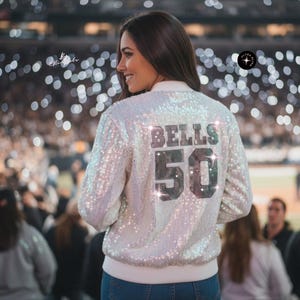 Könnte beinhalten: Eine weiße Pailletten-Bomberjacke mit dem Schriftzug "BELLS" und der Zahl "50" in dunkler, reflektierender Schrift auf dem Rücken. Die Jacke hat einen gerippten weißen Kragen und Saum, lange Ärmel und eine schimmernde, irisierende Oberfläche.