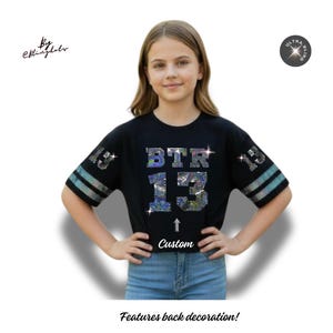 May include: Black short-sleeved shirt with the text "BTR 13" in sparkling rhinestones. The shirt has silver stripes on the sleeves and the word "Custom" below the number 13. The shirt is worn by a person.