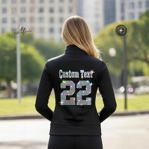May include: A black long-sleeved zip-up jacket featuring the text Custom Text and the number 22 on the back, both rendered in sparkling silver rhinestones. The jacket has a high collar and a fitted silhouette, showcasing a reflective crystal finish.