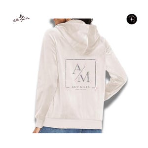 May include: A cream-colored zip-up hoodie featuring a back graphic with a square border, the letters AM, and the text AMY MILES REAL ESTATE. The design is embellished with small, sparkling rhinestones throughout the logo and border elements.