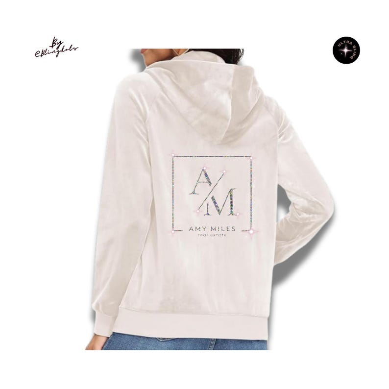 May include: A cream-colored zip-up hoodie featuring a back graphic with a square border, the letters AM, and the text AMY MILES REAL ESTATE. The design is embellished with small, sparkling rhinestones throughout the logo and border elements.