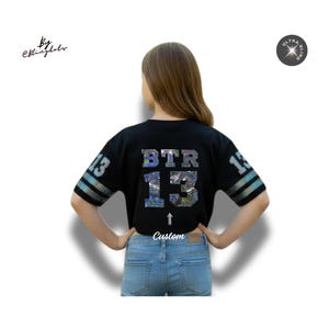 May include: Black crop top with the letters "BTR" and the number "13" in a shimmering, iridescent design. The sleeves have a striped pattern. The word "Custom" is below the number. The shirt is worn with blue jeans.