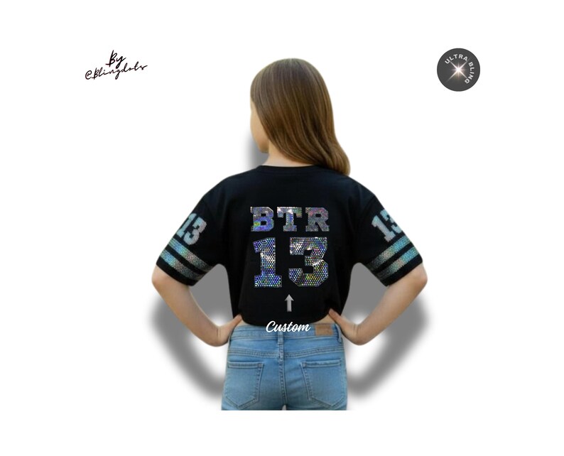May include: Black crop top with the letters "BTR" and the number "13" in a shimmering, iridescent design. The sleeves have a striped pattern. The word "Custom" is below the number. The shirt is worn with blue jeans.