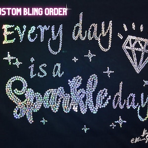 May include: A black t-shirt with the text "Every day is a sparkle day" in rhinestones. The text is surrounded by small stars and a large diamond shape also made of rhinestones. The bottom right corner of the shirt has the text "By @blingdots".