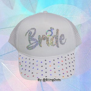 May include: White trucker hat with the word "Bride" spelled out in iridescent rhinestones. The brim is decorated with iridescent rhinestones. The hat has a white mesh back.