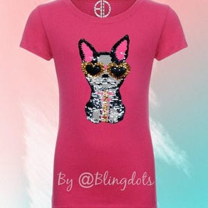 May include: Pink short-sleeved t-shirt with a sequined image of a dog wearing sunglasses. The text "By @Blingdots" is printed on the bottom of the shirt.