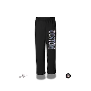 May include: Black sweatpants featuring an elastic waistband and the word CUSTOM written vertically down the left leg in shimmering, multi-colored rhinestones. The text is oriented from the top down in a bold, sans-serif font style.
