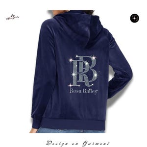 May include: A navy blue velour zip-up hoodie featuring a hood and long sleeves. The back displays a large, sparkling rhinestone monogram of the letters RB, with the name Rosa Bailey printed in white lettering directly underneath the monogram.
