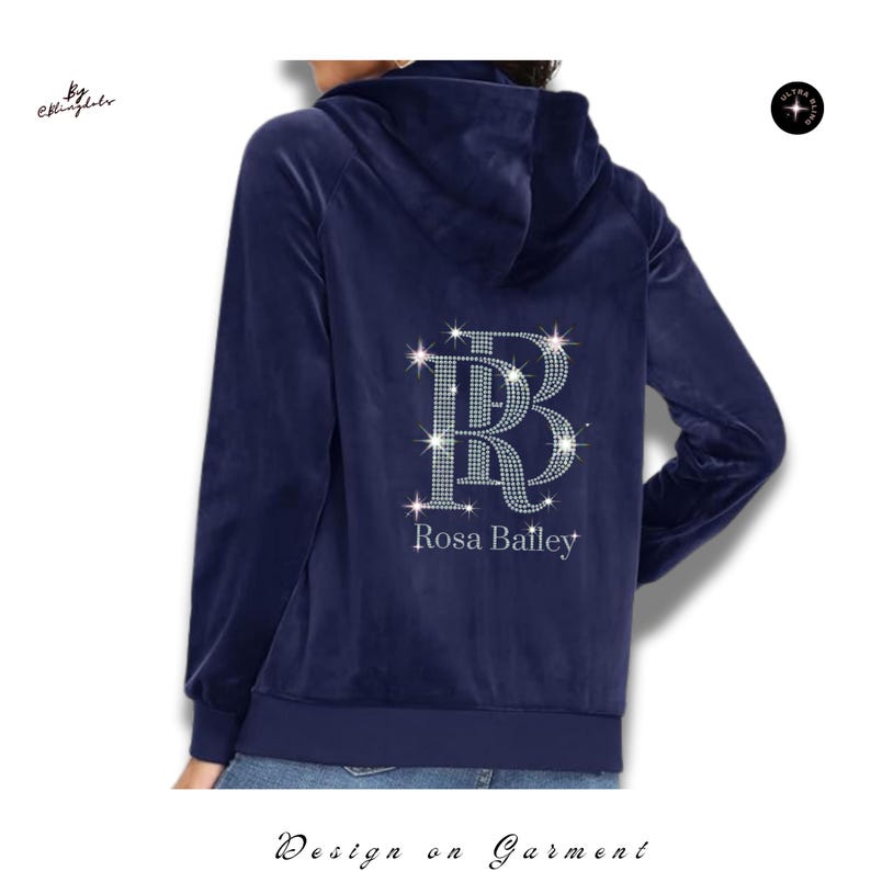 May include: A navy blue velour zip-up hoodie featuring a hood and long sleeves. The back displays a large, sparkling rhinestone monogram of the letters RB, with the name Rosa Bailey printed in white lettering directly underneath the monogram.