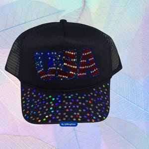May include: A black trucker hat with a mesh back. The front of the hat features a rhinestone design of the American flag with the word "USA" in red, white, and blue.