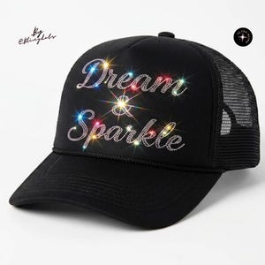 May include: A black trucker hat featuring the text Dream & Sparkle in clear rhinestones on the front panel. The cap has a curved brim, a mesh back, and a small circular logo in the top right corner with the text Ultra Bling and a star icon.