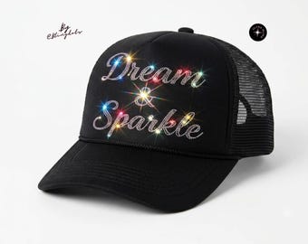 Op de afbeelding: Een zwarte truckerpet met de tekst Dream & Sparkle in doorzichtige strass-steentjes op het voorpaneel. De pet heeft een gebogen klep, een mesh achterkant en een klein rond logo in de rechterbovenhoek met de tekst Ultra Bling en een stericoon.