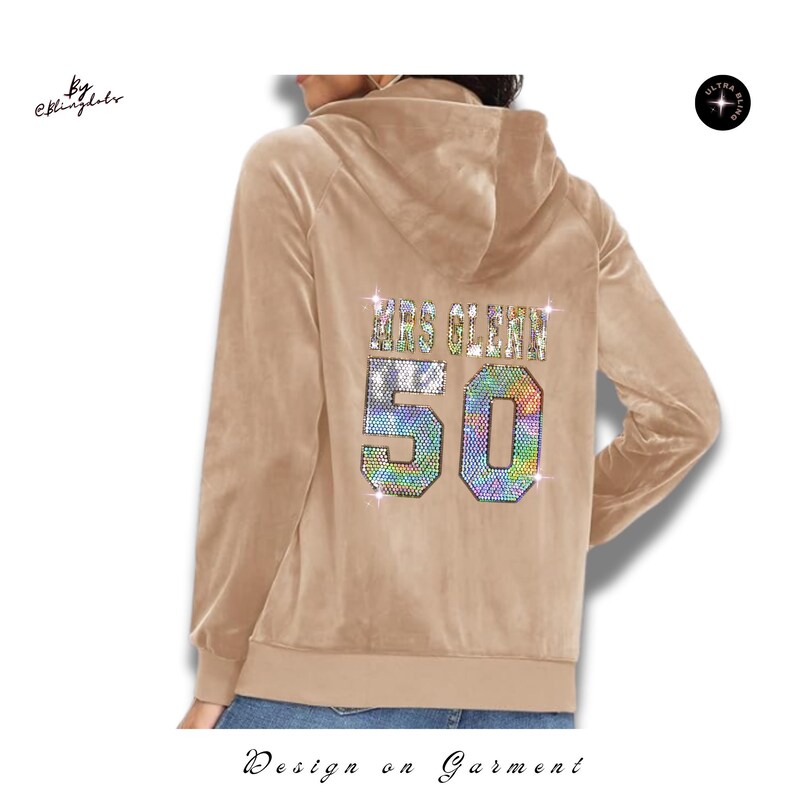May include: A beige velour zip-up hoodie featuring a hood and long sleeves. The back displays the text MRS GLENN above the large number 50, both rendered in multicolored, sparkling rhinestones that create a vibrant, reflective effect.