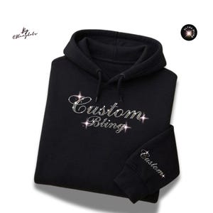 May include: A black hooded sweatshirt featuring the text Custom Bling in sparkling rhinestones on the chest and the word Custom in rhinestones on the sleeve cuff. The design includes star-shaped light reflections around the lettering.