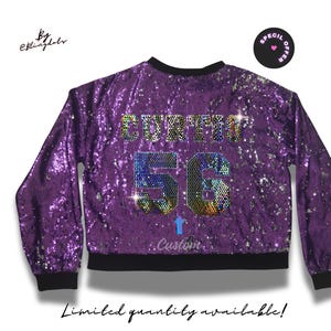 Może przedstawiać: Fioletowa bomberka z cekinów z imieniem "Curtis" i numerem "56" w tęczowych kryształach. Kurtka ma czarny, prążkowany kołnierz, mankiety i dół.