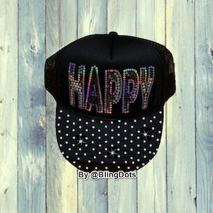 Peut inclure: Casquette de camionneur noire avec le mot "HAPPY" en strass irisés. La visière est noire avec des pois blancs. La casquette a un dos en filet et une fermeture à pression.