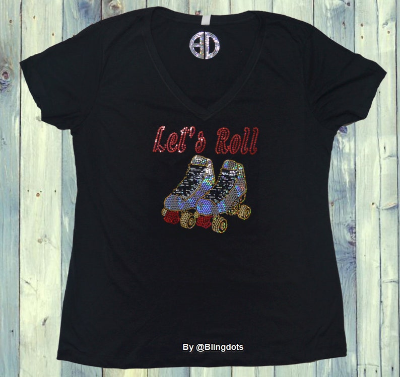 Puede incluir: Camiseta negra con un dise&ntilde;o de strass rojo que dice "Let's Roll" con un par de patines debajo. El dise&ntilde;o es brillante y llamativo.