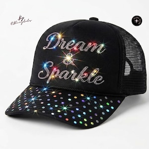 Puede incluir: Una gorra tipo camionero negra con el texto Dream & Sparkle en pedrería plateada en el panel frontal. La visera está decorada con un patrón de puntos iridiscentes multicolores. Se observa un pequeño logotipo circular con el texto Ultra Bling.