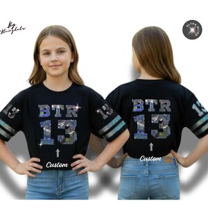 May include: Black cropped t-shirt with the text "BTR 13" in sparkling, iridescent rhinestones. The sleeves have silver and blue stripes and the number "13". The word "Custom" is below the number 13.