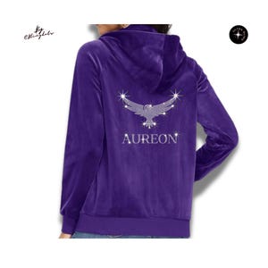 May include: A deep purple velour zip-up hoodie featuring a hood and long sleeves. The back displays a large, sparkling rhinestone graphic of an eagle with wings spread wide, positioned above the text AUREON in matching rhinestone lettering.