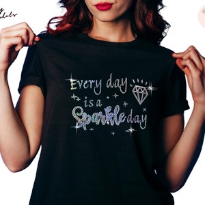 Könnte beinhalten: Schwarzes T-Shirt mit dem Schriftzug "Every day is a Sparkle day" aus glitzernden Strasssteinen. Das Design beinhaltet einen Diamanten und Sterne. Das Shirt hat einen Rundhalsausschnitt.