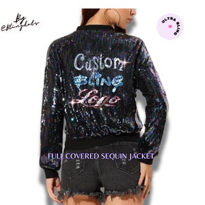 Puede incluir: Una chaqueta bomber negra de lentejuelas con el texto "Custom Bling Logo" en lentejuelas de colores. La chaqueta está completamente cubierta de lentejuelas y tiene una cremallera.