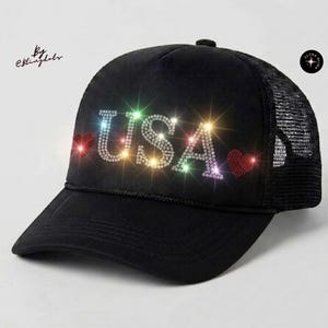 May include: A black trucker hat featuring the text USA in clear rhinestones, flanked by two red rhinestone hearts. The front panel is solid black, while the back panels are made of black mesh. The design sparkles with multi-colored light reflections.