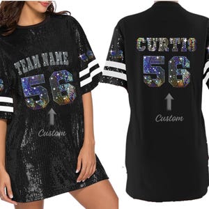 Puede incluir: Vestido de jersey negro con lentejuelas y rayas blancas y plateadas en las mangas. La parte delantera del vestido tiene el texto "TEAM NAME" y el n&uacute;mero "56" en pedrer&iacute;a iridiscente. La parte trasera del vestido tiene el nombre "CURTIS" y el n&uacute;mero "56" en pedrer&iacute;a iridiscente.