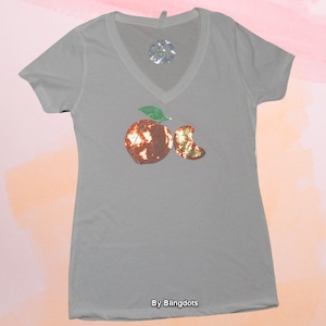 May include: White v-neck t-shirt with a peach design made of rhinestones. The peach is orange and has a green leaf. The text "By Blingdots" is at the bottom of the shirt.