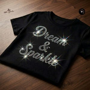Puede incluir: Una camiseta negra de manga corta con el texto Dream & Sparkle formado por brillantes transparentes y reflectantes. Las letras están dispuestas en tres líneas en el centro del pecho, creando un efecto brillante y centelleante sobre la tela.