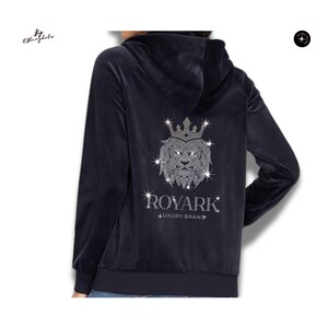 May include: A dark navy blue velour zip-up hoodie featuring a large, sparkling rhinestone lion head graphic wearing a crown on the back. Below the lion, the text reads ROYARK LUXURY BRAND in silver rhinestones. The hoodie has long sleeves and a hood.