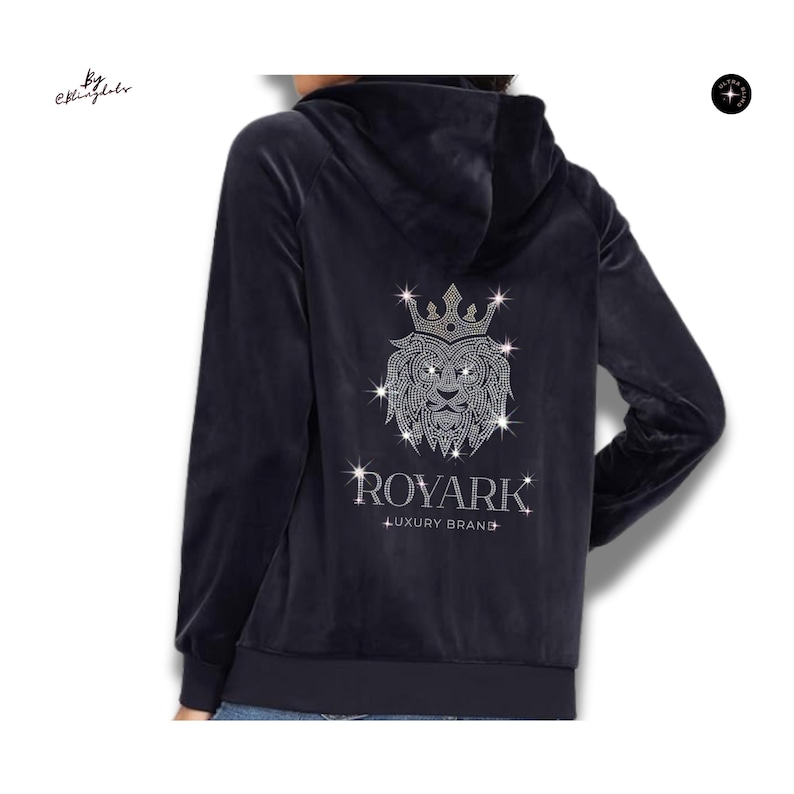 May include: A dark navy blue velour zip-up hoodie featuring a large, sparkling rhinestone lion head graphic wearing a crown on the back. Below the lion, the text reads ROYARK LUXURY BRAND in silver rhinestones. The hoodie has long sleeves and a hood.