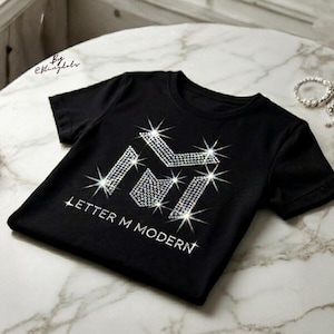 May include: A black short-sleeved t-shirt featuring a large letter M graphic constructed from clear, sparkling rhinestones. Below the letter M, the text "LETTER M MODERN" is also written in clear rhinestones. The design creates a starburst effect.
