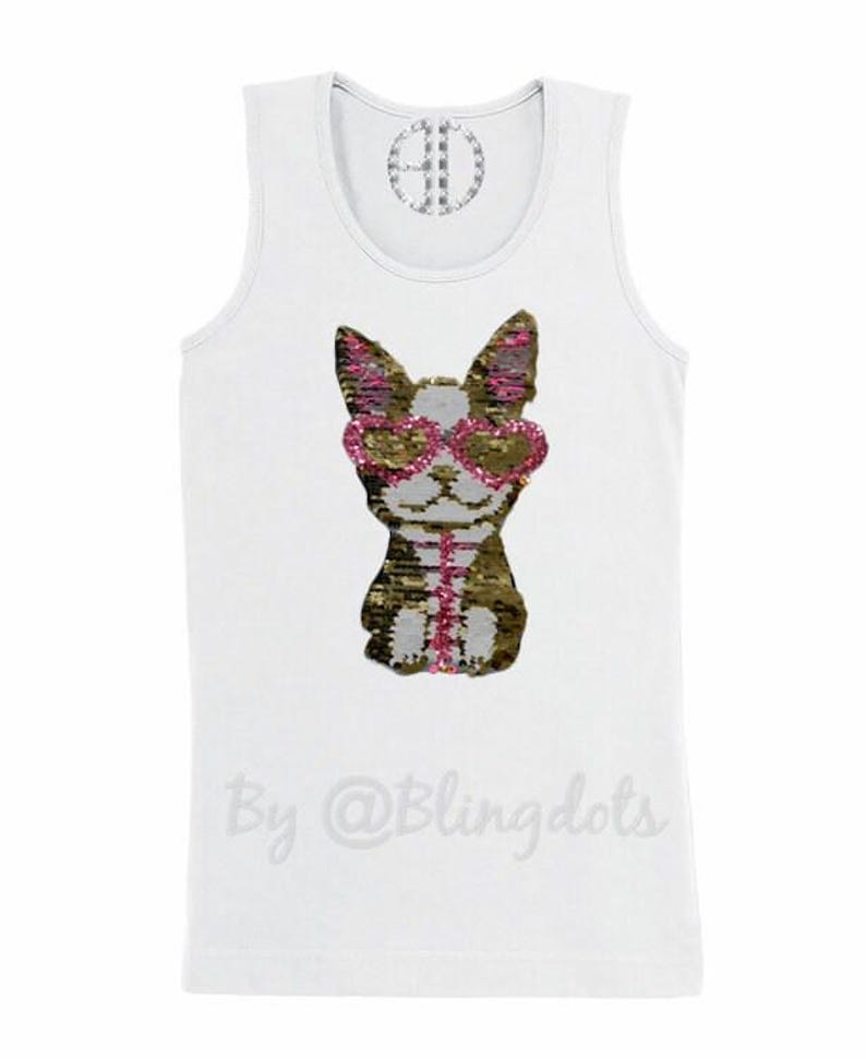 May include: White tank top with a sequined dog wearing pink heart-shaped sunglasses. The dog is brown and gold with pink accents. The text "By @Blingdots" is printed on the bottom of the shirt.