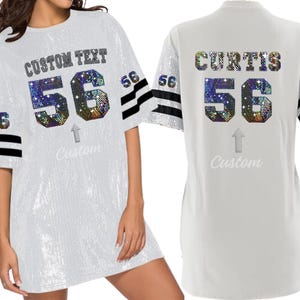 Puede incluir: Vestido de jersey blanco con rayas blancas y negras en las mangas. La parte delantera del vestido tiene el texto "CUSTOM TEXT" y el n&uacute;mero "56" en pedrer&iacute;a iridiscente. La parte trasera del vestido tiene el nombre "CURTIS" y el n&uacute;mero "56" en pedrer&iacute;a iridiscente.